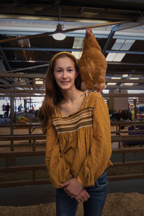 A young girl stands with a bird on her left shoulder. Her brownish shirt and hair band match the color of the bird.