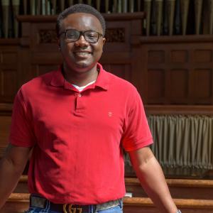 A smiling young man wearing a red shirt stands between church pews.