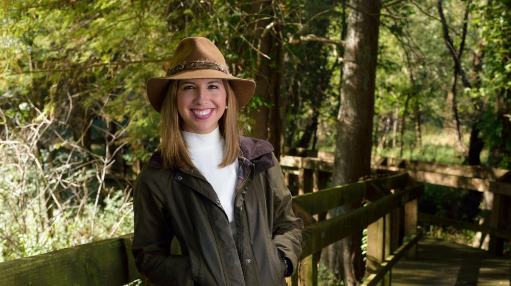 A young woman with brown hair wearing a tan hat with a green jacket and khaki pants leans against a wooden railing.