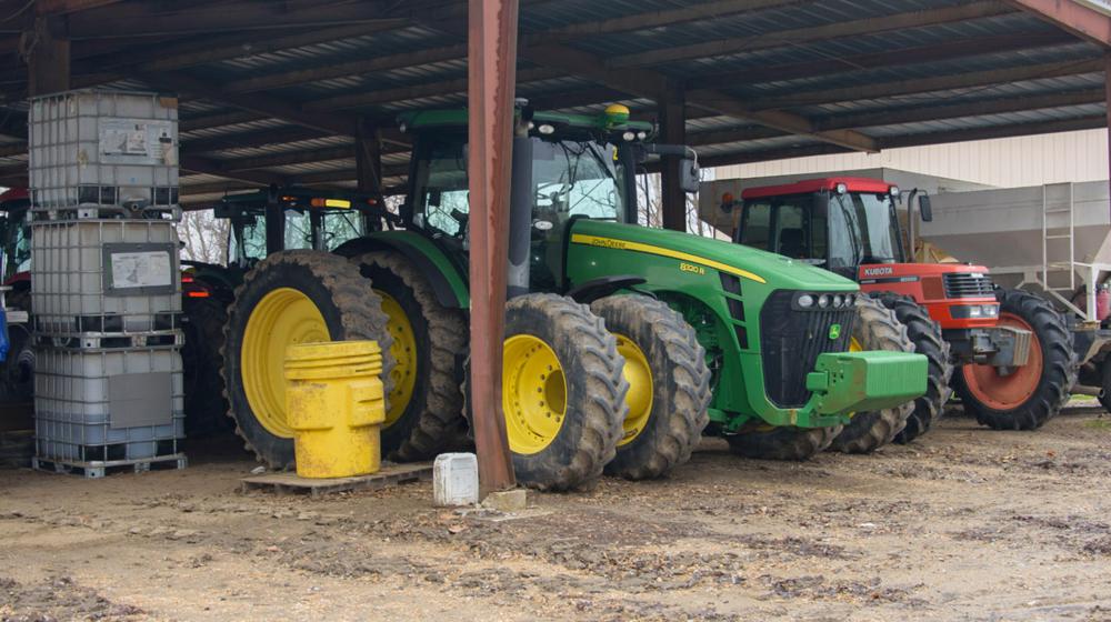 A green tractor, flanked on the right by a smaller red one, stands beneath a metal roof.