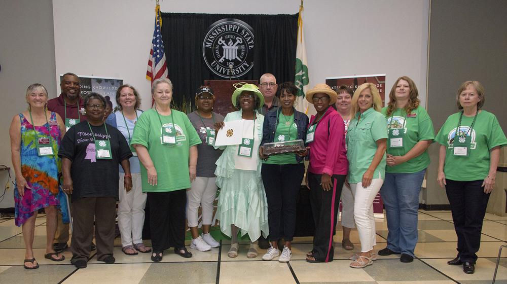 Mississippi Volunteer Leaders Association A group of 14 men and women stand on either side of a woman wearing a bright green hat and holding up an award.