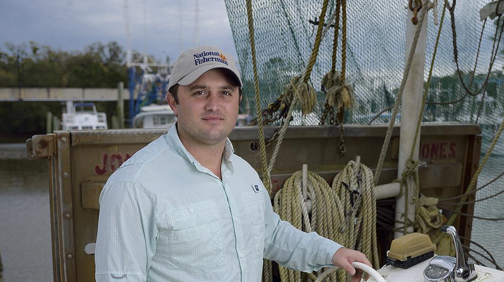 Ryan Bradley, Commercial Angler A man wearing a blue collared shirt stands in a boat with his hand on an old white steering wheel.