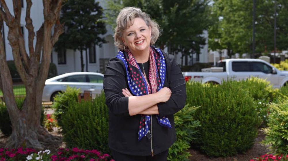 A woman, wearing all black clothes and a red, white, and blue scarf, smiling in front of flower bushes with her arms crossed.