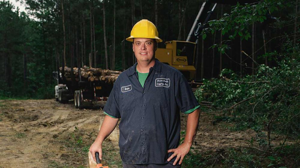 A man wearing a yellow hard hat and holding a chain saw stands in front of a trailer holding harvested timber.