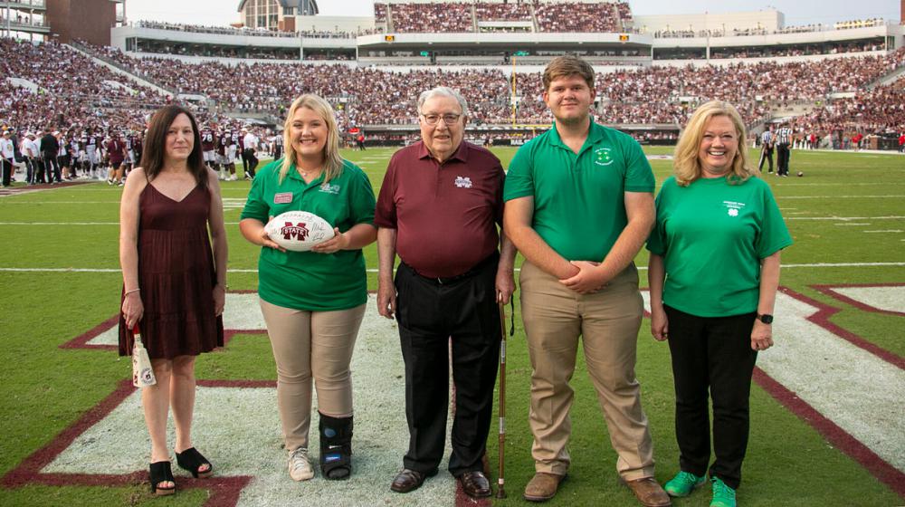 A group of five men and women standing on the MSU football field during a game day. 
