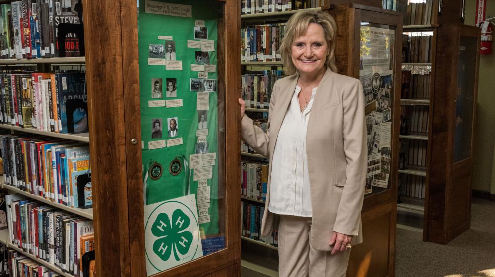 A woman stands in front of library book shelves beside a tall, green 4-H display.