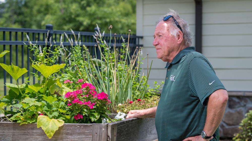A man, standing beside a wooden table filled with plants, looking into the distance.