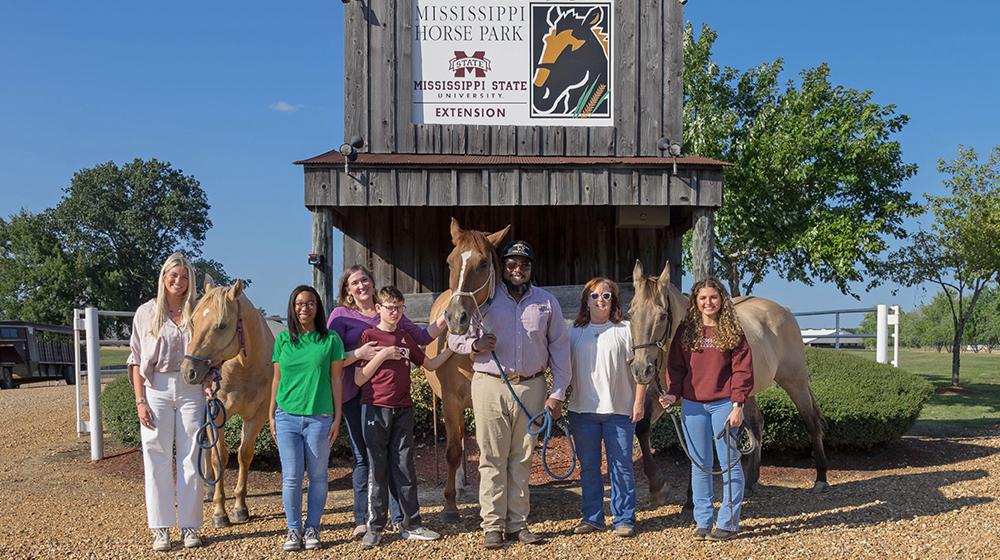 A group of men and women standing in front of a barn and alongside three horses.