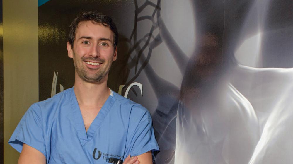 A man in blue scrubs with crossed arms leans against a wall.