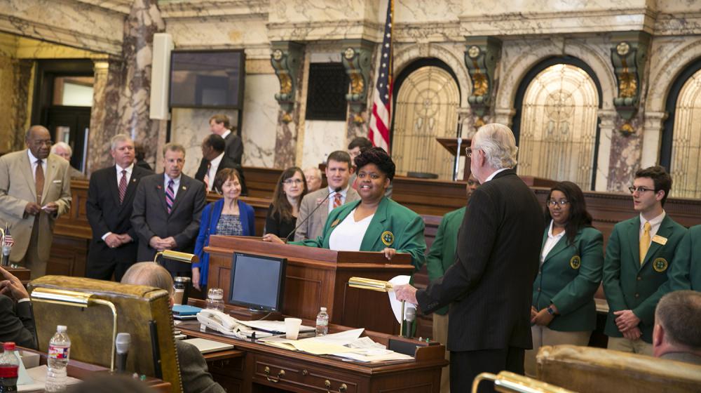 Standing Ovation A young woman wearing a dark green blazer stands behind a podium while several people stand looking at her.