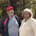 A couple, a man with a black and red jacket and a woman with a tan cardigan, stands next to a wooden railing located outside with trees and water in the background.