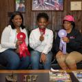 A young female dressed in a white polo and blue jeans smiles and holds up the second-place ribbon around her neck. On the left is an adult woman dressed in a white sheer top with a white vest and blue jeans and is holding a bigger red ribbon. On the right is an older woman wearing a pink AKA hat and a black long-sleeved AKA T-shirt with brown slacks, and she holds up a purple ribbon.