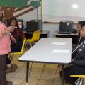 A woman, leftmost, wearing a coral blouse and brown slacks, points and talks to two teens sitting in yellow chairs at a gray table.