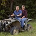 A man wearing a blue shirt and a woman wearing a maroon shirt sitting on a four-wheeler in tall green grass in front of dark green trees.