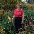 A woman wearing a red collared shirt standing in tall green grass and flowers. She holds a shovel in her right hand, which rests in front of a metal butterfly garden decoration.