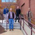 Three men and one woman standing, spaced out in front of a red brick building.