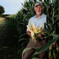A man holding three ears of corn stands in a corn field.