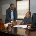 A standing Black man points to a paper on a desk that a seated white man with a pen also points to.