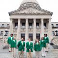 Seven teenagers wearing green blazers and tan slacks form a U standing on steps in front of six gray columns.