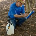 A man kneels on the ground beside a pump sprayer to read the label on the back of a pesticide jar.