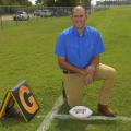 A man in a blue shirt kneels on a chalk-lined football field beside a goal line marker. A white Mississippi State football rests on the ground beside his knee.