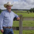 Man wearing a cowboy hat standing against a fence in front of a field with cows grazing.