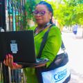 Woman in a green dress with a bag around her shoulder stands in front of a gate holding a laptop computer.