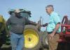 Two men facing each other in conversation and standing beside a tractor and equipment with a clear, blue sky overhead.