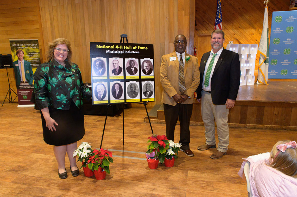 Three people standing and smiling beside a poster board showing pictures of the National 4-H Hall of Fame inductees from Mississippi.