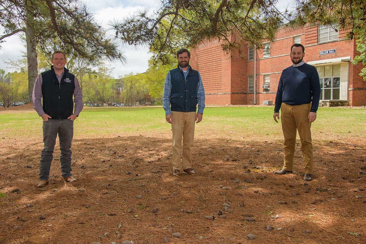 Three smiling men standing several feet apart from each other under the shade of a tree.