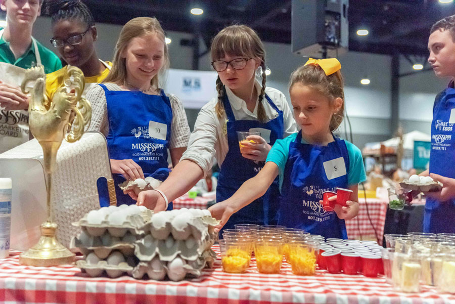 Three girls wearing blue aprons reach toward a tray of eggs.