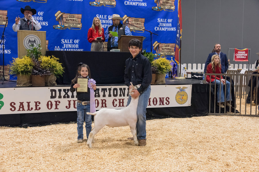 A girl holds a certificate and purple ribbon beside a young man holding a goat’s head.