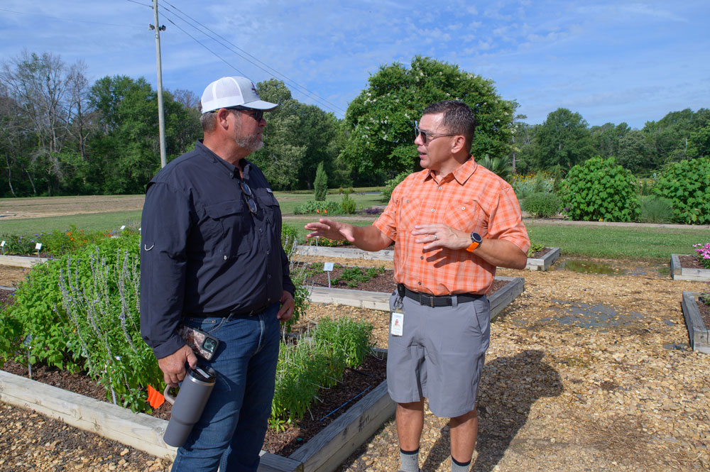 A man in an orange shirt talking with a man wearing a baseball hat and standing in a raised bed garden.