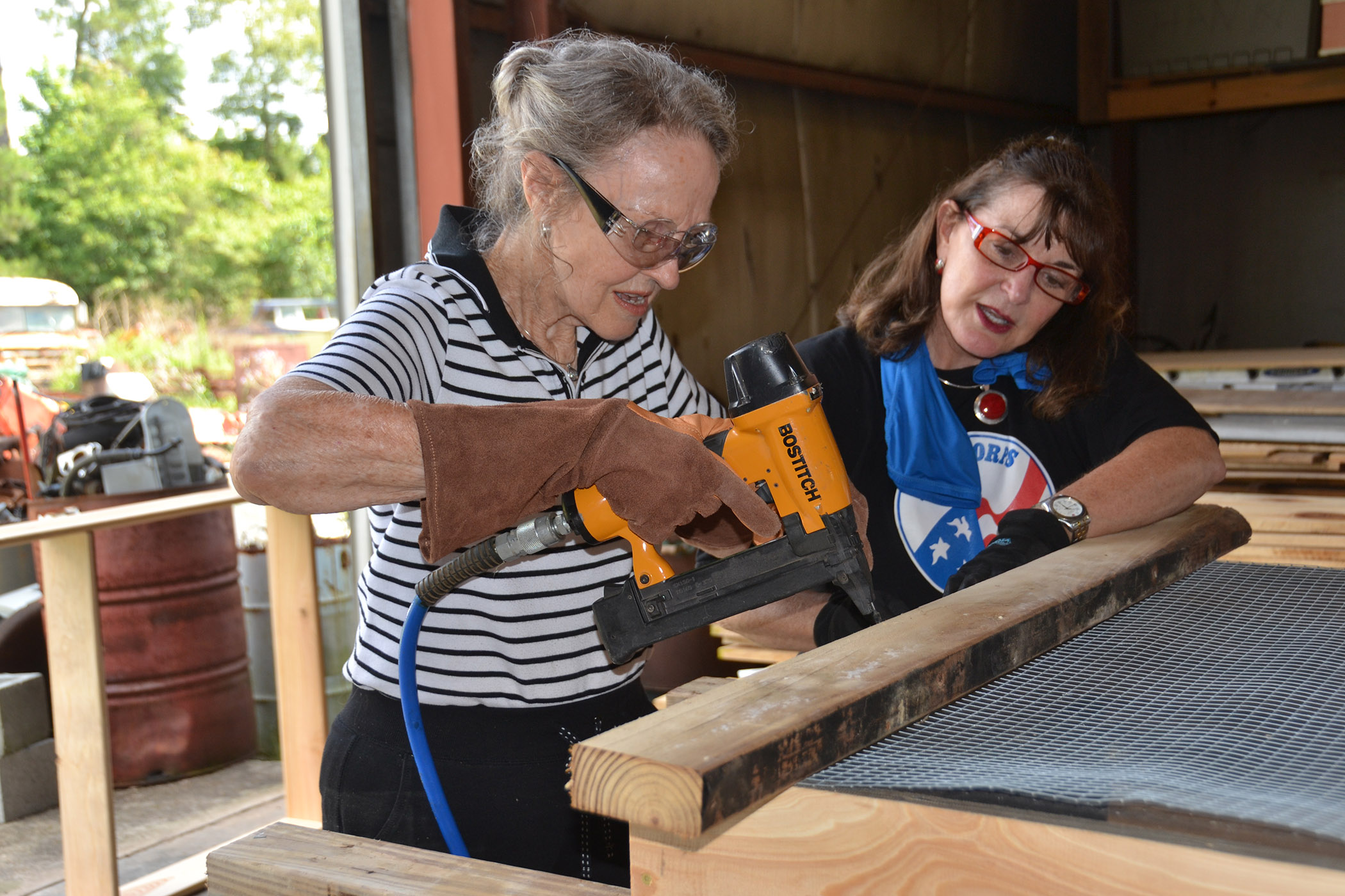 Alt-text: A woman stables hardware cloth to the wooden frame of a salad table while another woman helps hold the cloth in place.  