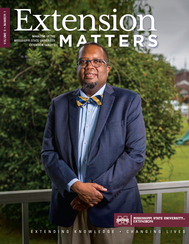 A Black man with glasses wearing a blue and yellow striped tie, a blue dress jacket, and khakis, standing on a deck in front of a tree.