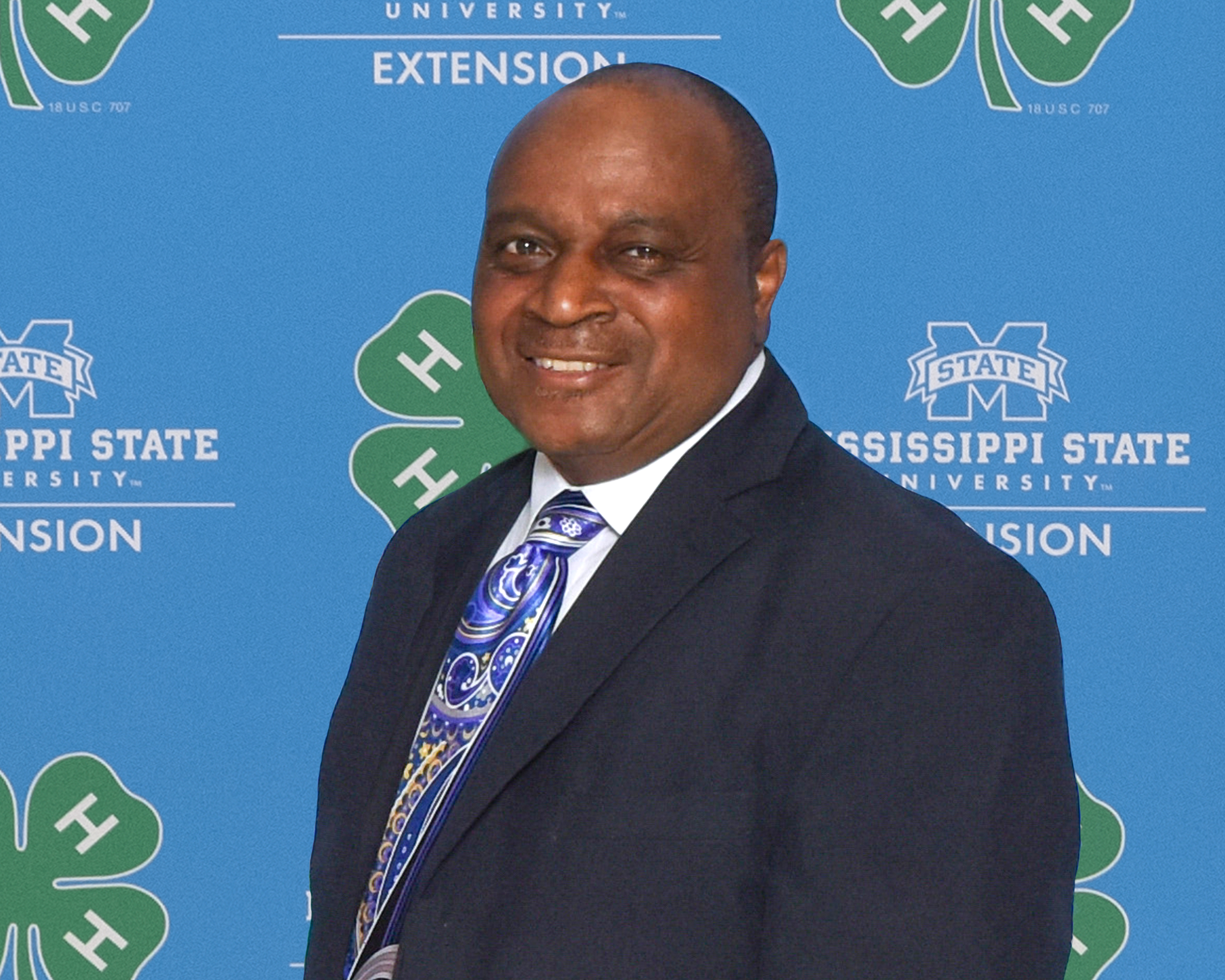 A smiling Black man wearing a suit stands in front of a 4-H background.