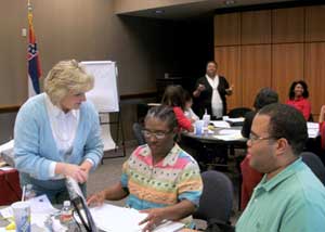 Standing woman in blue and white blouse talks to two seated people at a table.