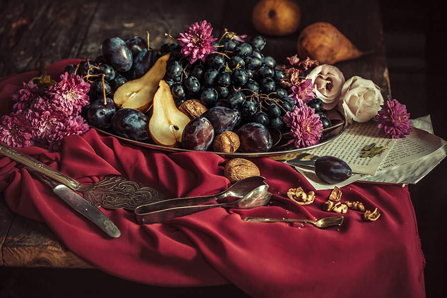 A bowl of fruit with dark violet, red-violet, and dark tan are on a red-violet table covering.