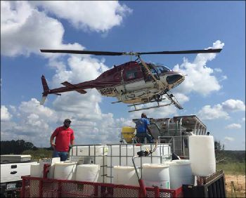 A helicopter is hovering over a refilling station with two workers on the ground. 