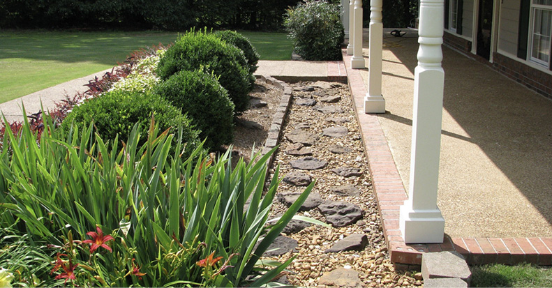 iew from a covered front porch showing a border area filled with ornamental gravel and stones and edged with pavers. This border separates the porch from a traditional ornamental bed with shrubs, flowering plants, and ornamental grasses.