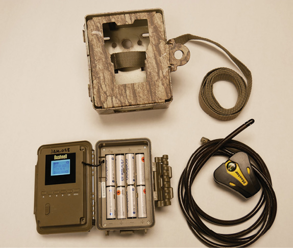 A layout of items including a camouflage security box, cable lock with cable, and Bushnell camera displaying its battery panel with eight installed rechargeable batteries.