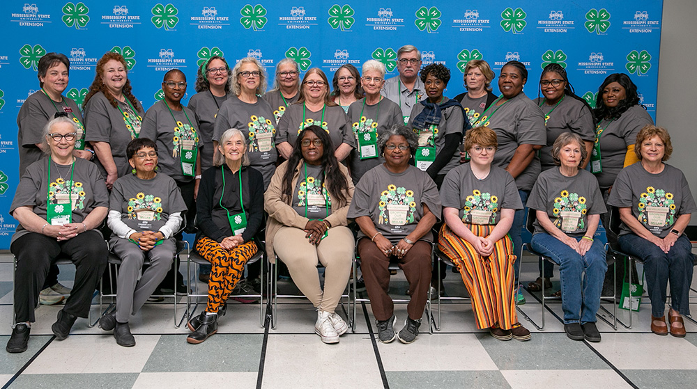 Two rows of 4-H volunteers, with the front row sitting down with hands in their laps and the back row standing, in front of a blue background