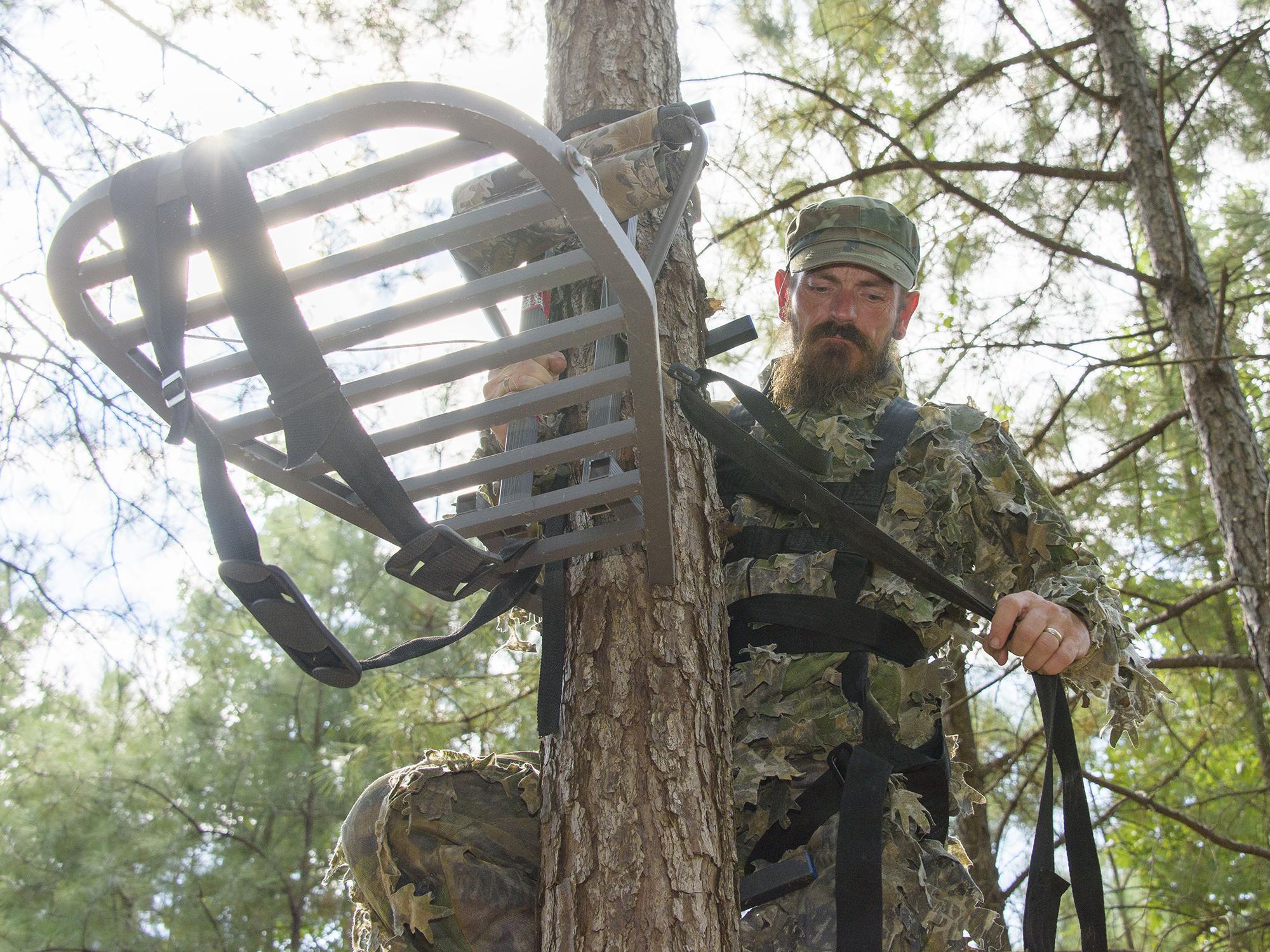 Hunter wearing camouflage secures a portable platform to the side of a tree.