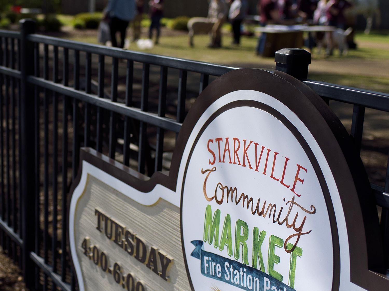 Shoppers can be seen browsing in the background of a farmers market sign.