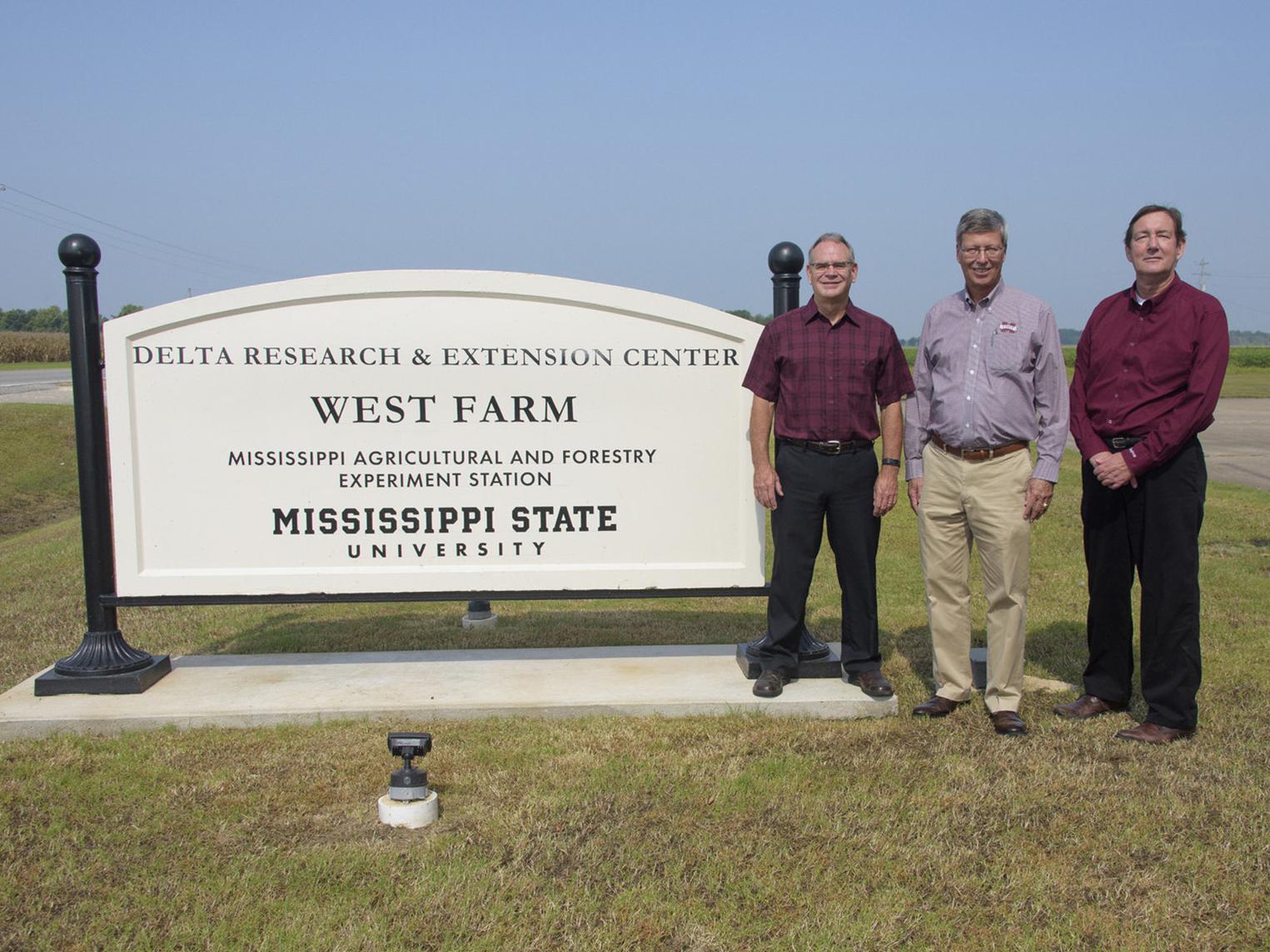 Three men stand beside a white sign on a cloudless day. The sign reads “Delta Research & Extension Center, West Farm, Mississippi Agricultural and Forestry Experiment Station, Mississippi State University.”