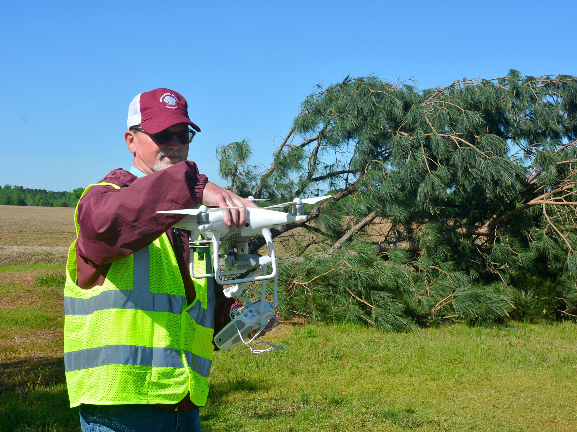 Man wearing a reflective safety vest looks at a white drone he is holding at shoulder height. A toppled pine tree and empty agricultural field are in the background.