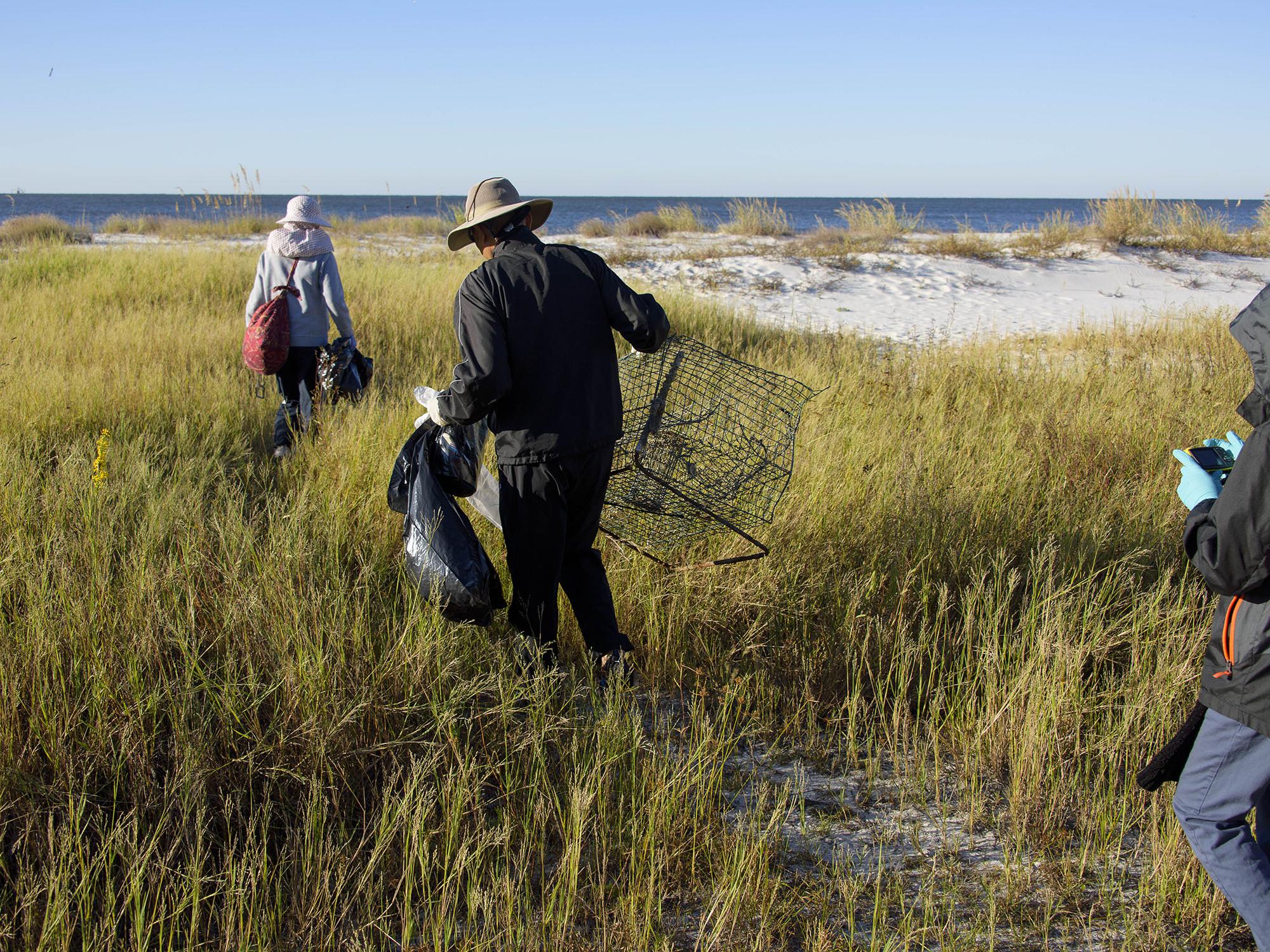 A man in the center of the photo is shown from the back wearing a bucket hat and black wind suit picking up trash in tall grass along a beach. Another person with a gray jacket and red backpack is in front of him with a trash bag, while another person in a black jacket with the hood up takes pictures. A blue sky and ocean water are in the background.