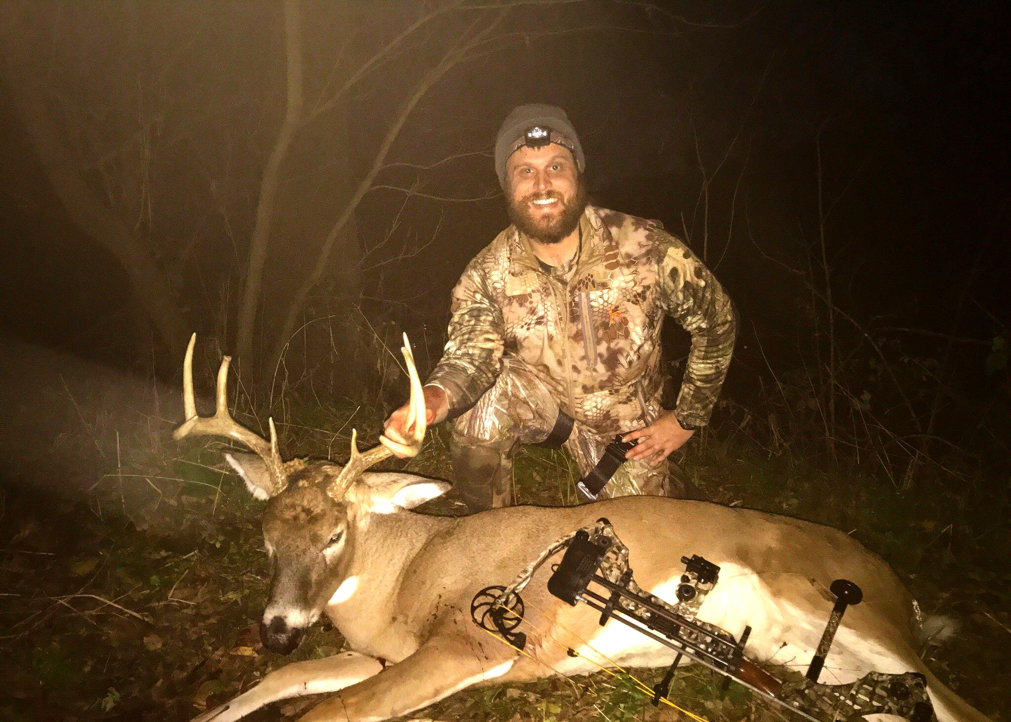 A man wearing hunting gear displays a deer he harvested with a bow and arrow.