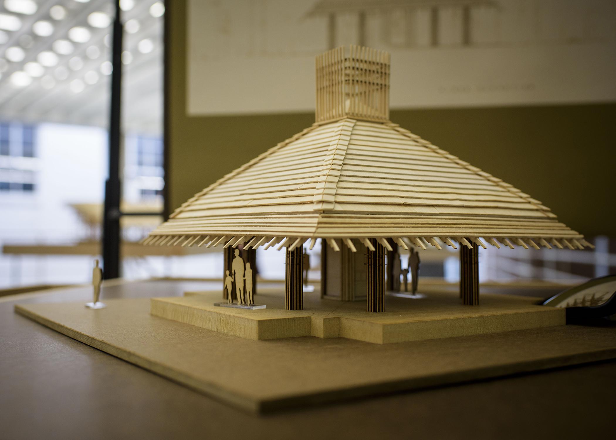 A wooden model of a building rests on a table in an exhibit hall.