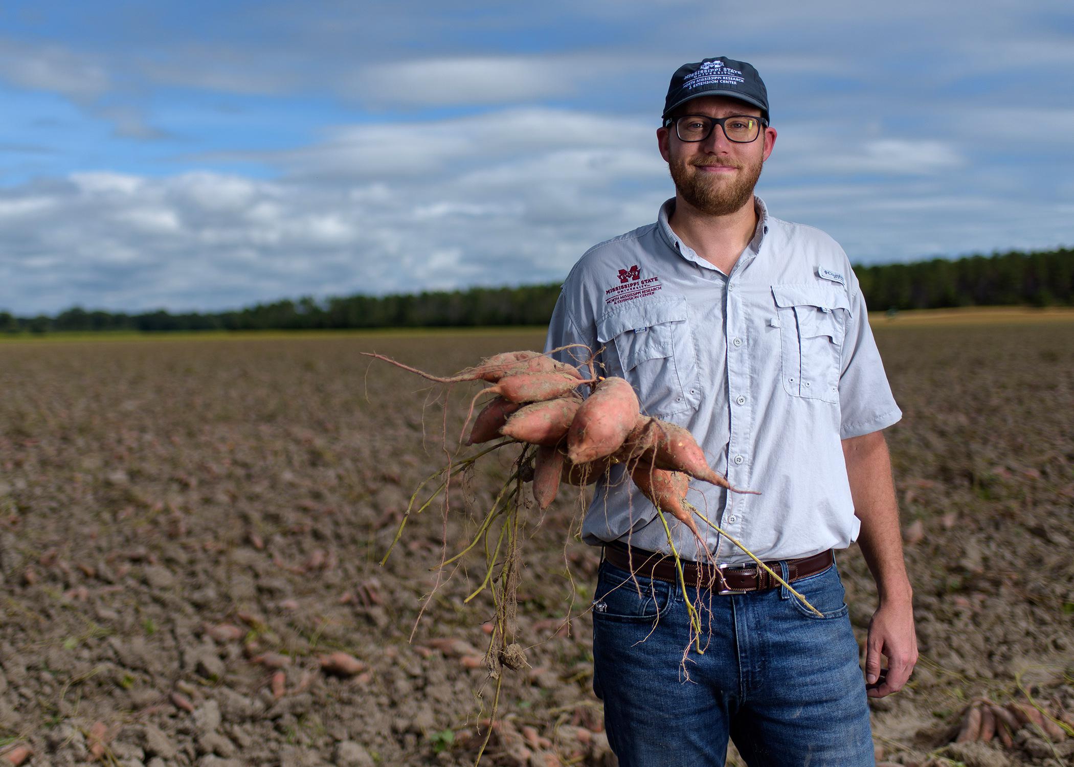 A man stands in a field with sweet potatoes in his hand.